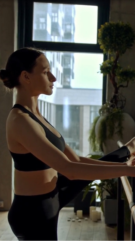 a woman performing ballet exercise on dance studio flooring utilizing a dance bar and a window and plants in the background